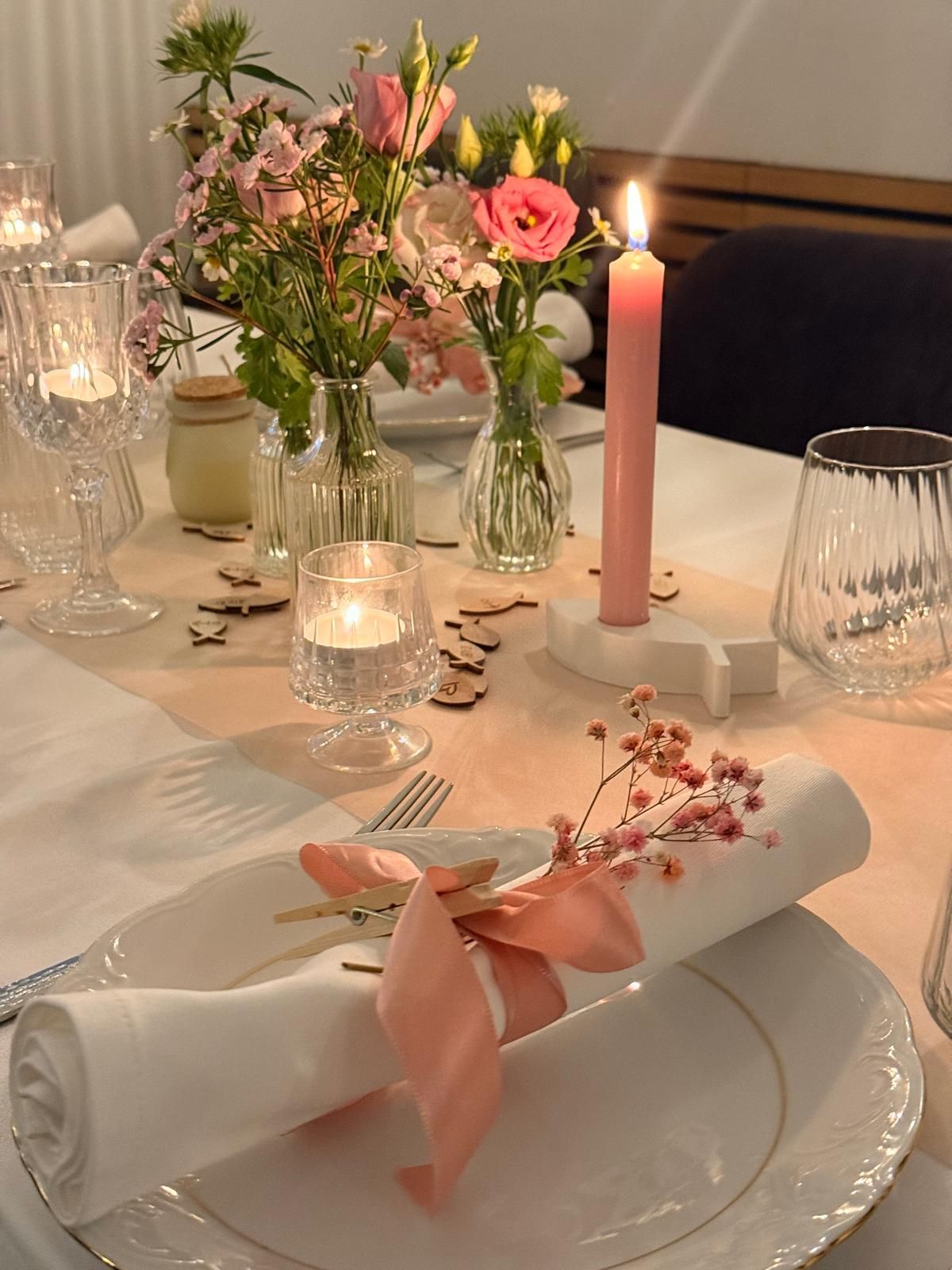 Elegant gedeckter Esstisch mit rosa Blumenarrangement, Kerze und gerollten Servietten mit rosa Band