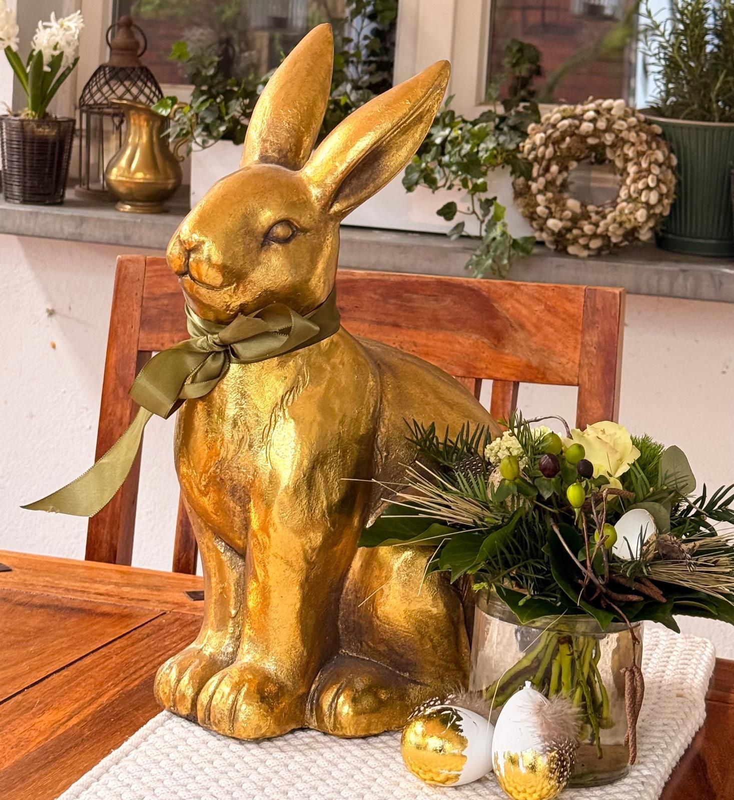 Goldene Hasenstatue mit grüner Schleife und Blumen auf einem Holztisch im Freien