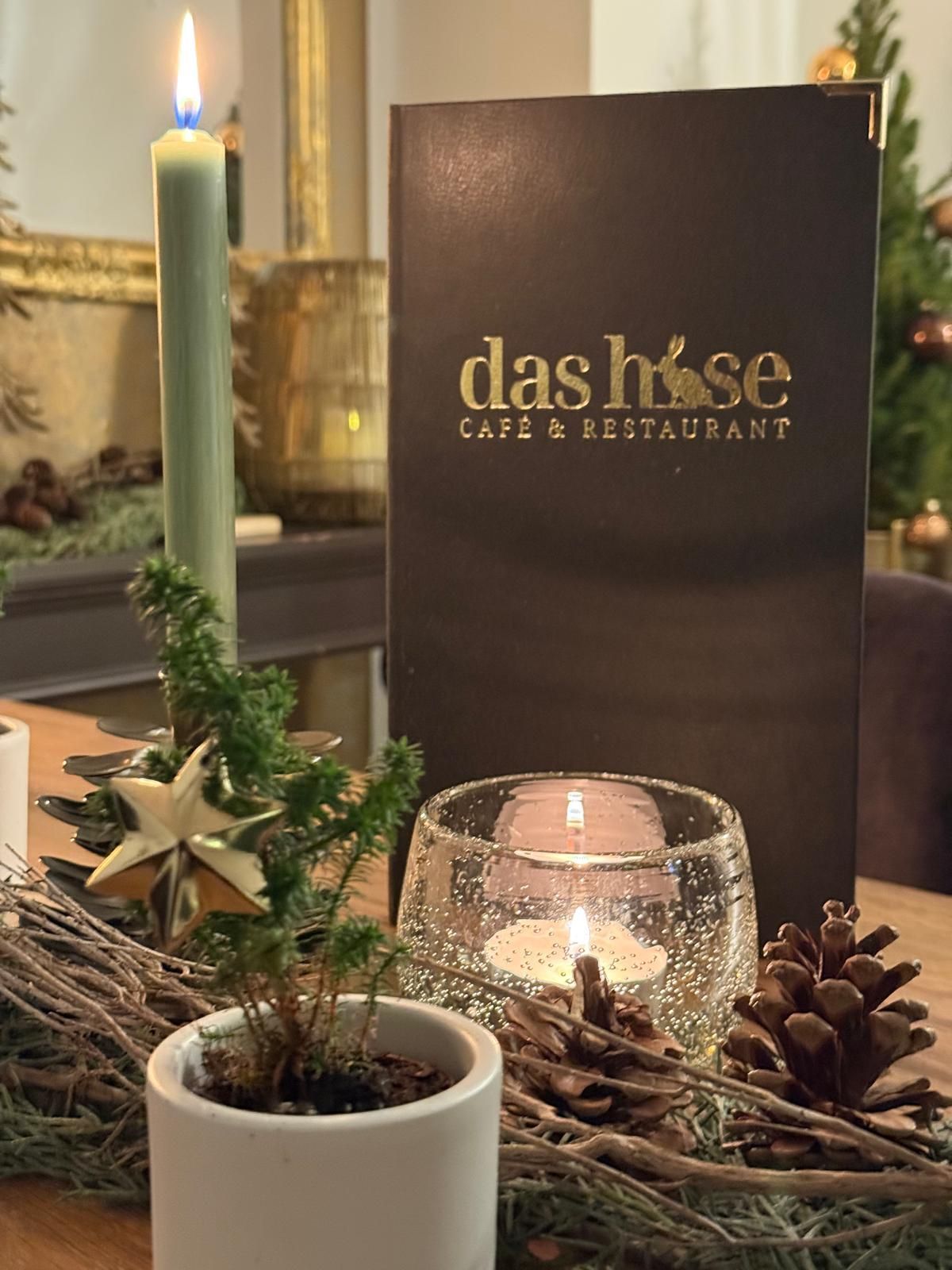 Tischdekoration im Kerzenschein mit immergrünem Zweig, Tannenzapfen und der Speisekarte des Das Hase Café & Restaurant im Hintergrund
