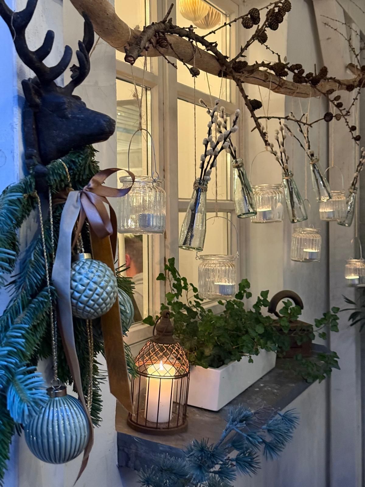 Dekoriertes Schaufenster mit hängenden Ornamenten, Topfpflanzen und warmem Kerzenlicht in einem gemütlichen Interieur