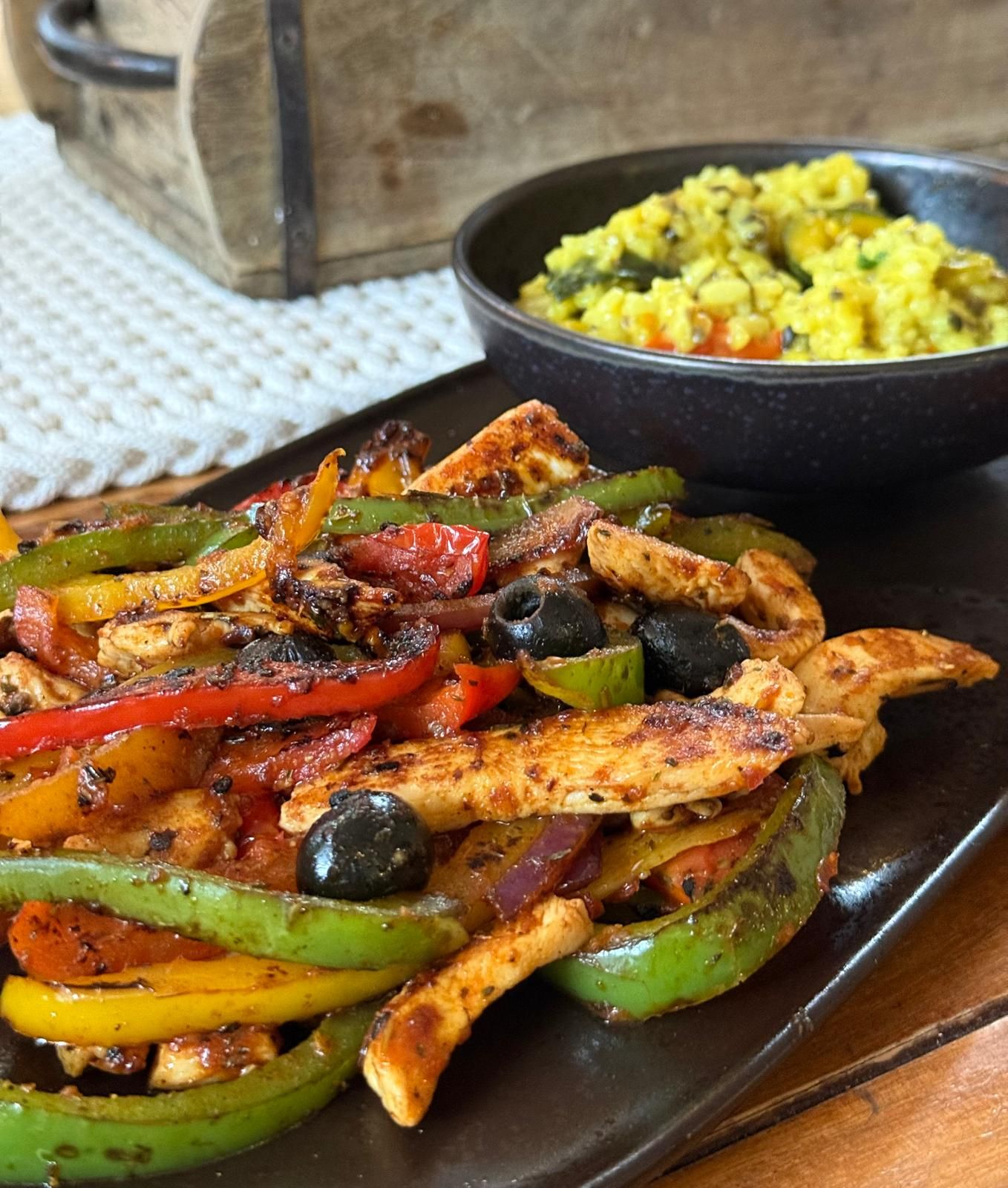 Eine brutzelnde Fajita-Platte mit Paprika und Zwiebeln, serviert neben einer Schüssel gelbem Reis auf einem Tisch.