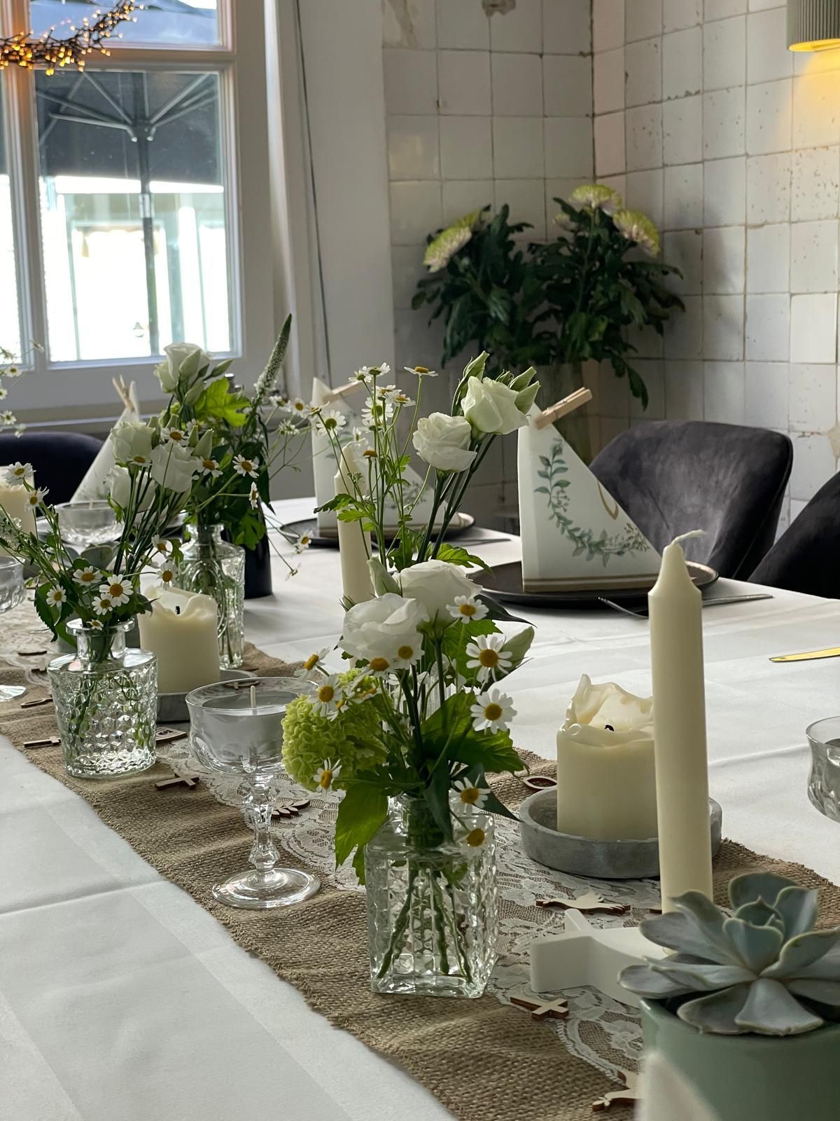 Ein weißes Blumengesteck als Tischdekoration auf einem gedeckten Esstisch mit Kerzen und Sukkulenten in einem hellen Raum