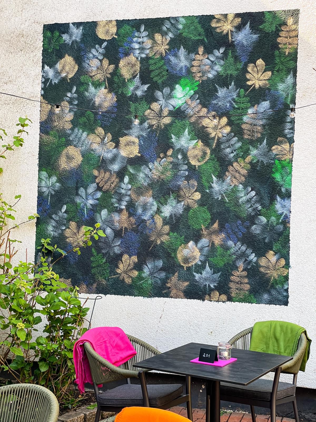 Terrasse im Freien mit floraler Wandkunst, einem kleinen schwarzen Tisch und bunten Stühlen mit rosa und grünen Decken.