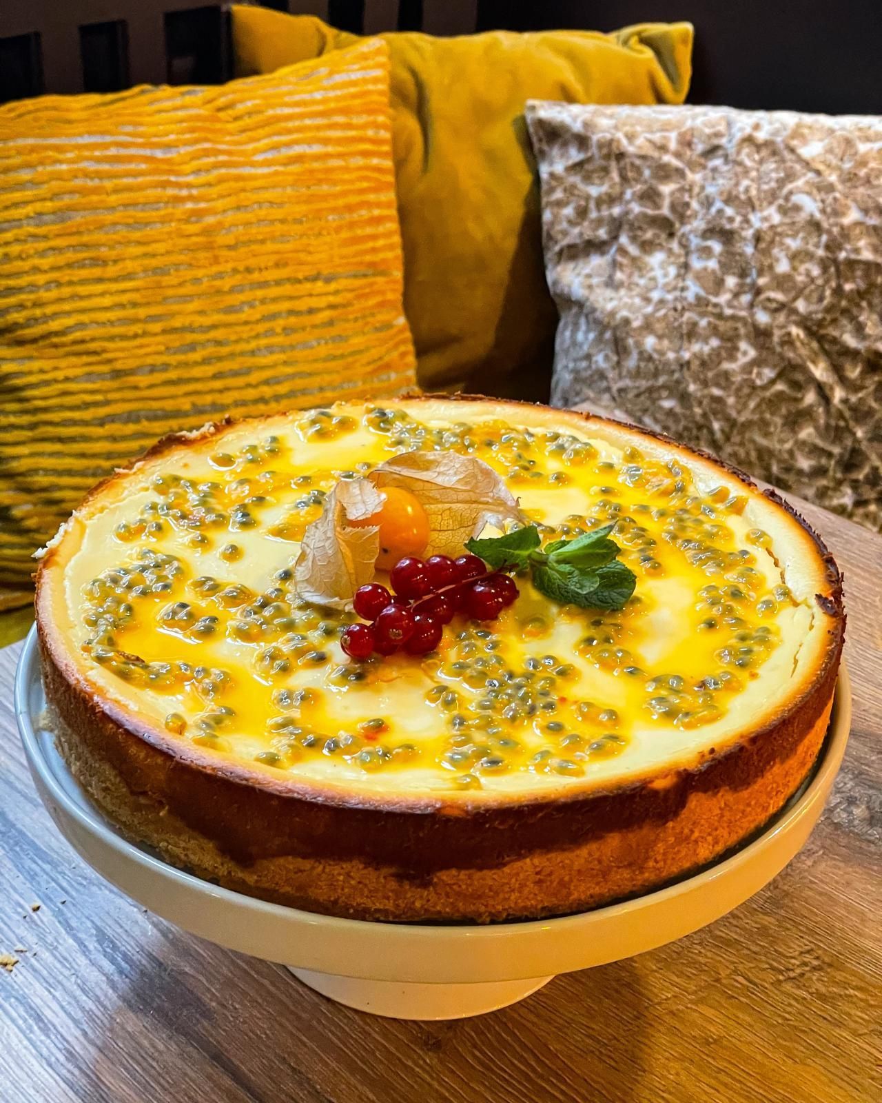 Ein mit Beeren garnierter Käsekuchen auf einem Teller, im Hintergrund ein gelbes Sofa und gemusterte Kissen.
