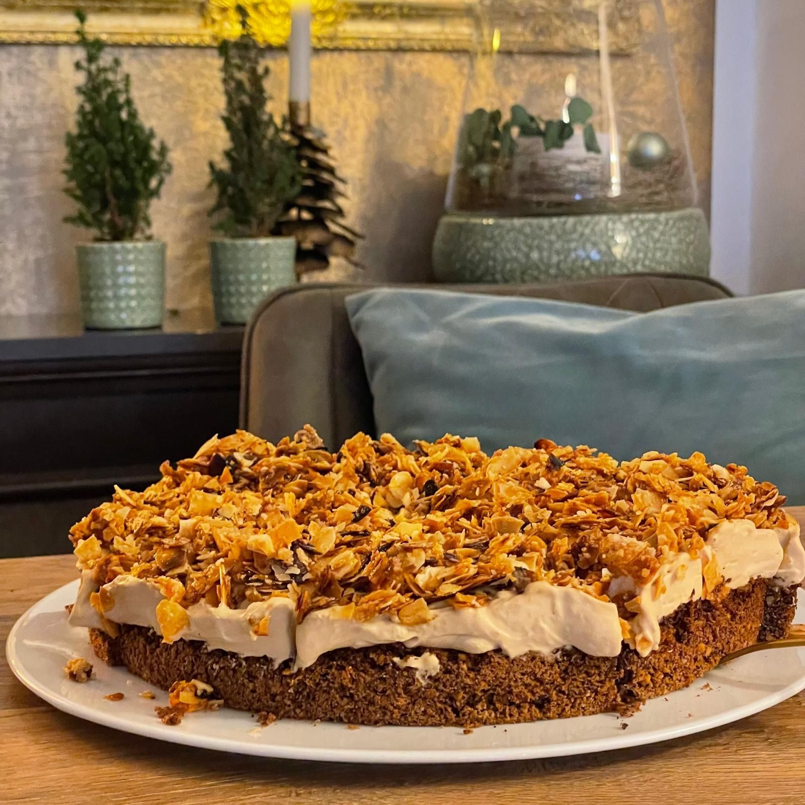 Ein Stück geschichtetes Dessert mit Streuseln auf einem weißen Teller, angerichtet auf einem Tisch in einem gemütlichen Wohnzimmer.