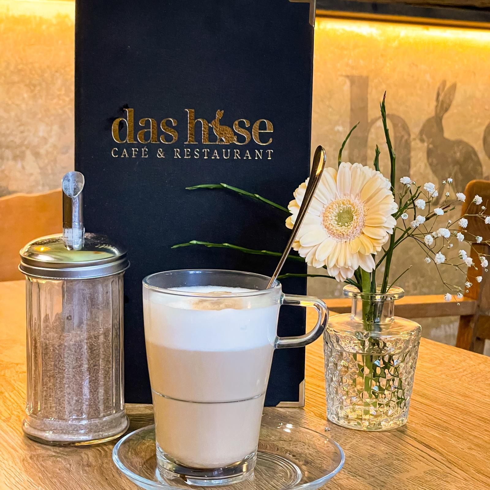 Café-Tisch mit Latte Macchiato, Zuckerdose, Blumen und dahinter eine schwarze Menütafel.