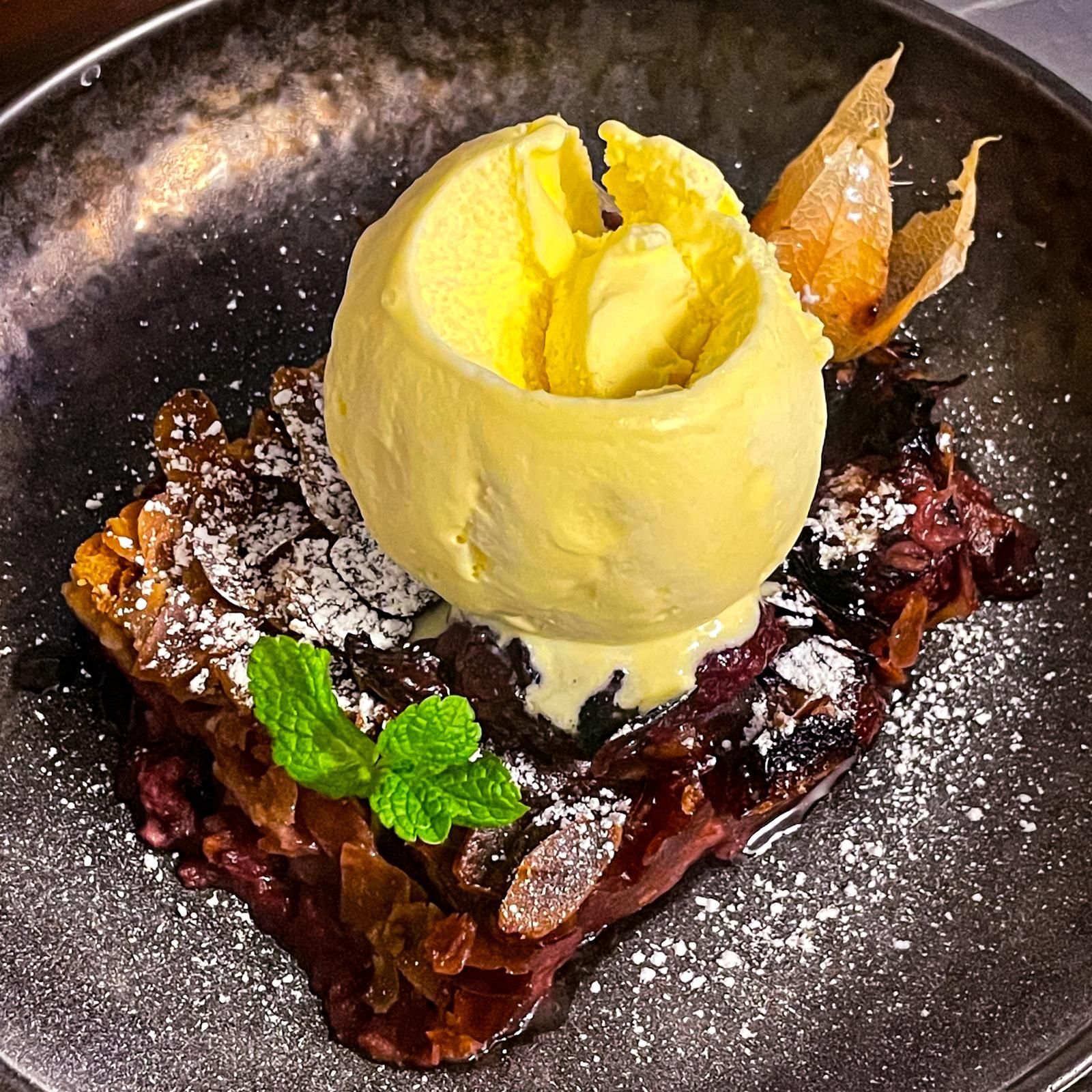 Eine Kugel gelbes Eis auf einem mit Puderzucker bestäubten Schokoladendessert in einer schwarzen Schüssel