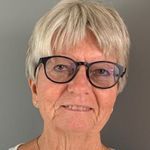 Smilende person med kort blondt hår og svarte briller mot en grå bakgrunn