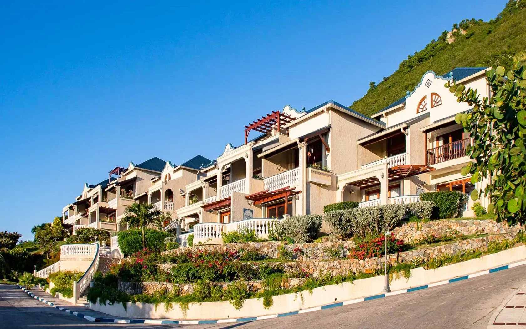 Row of light-colored buildings with balconies and landscaping, built into a hillside under a blue sky.
