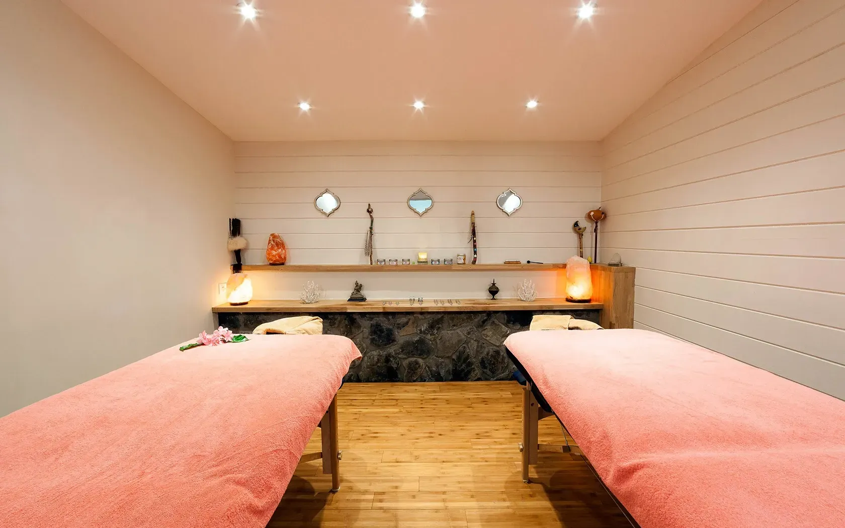 Two massage tables in a spa room, pink linens, wooden floors, white and wood-paneled walls, soft lighting.
