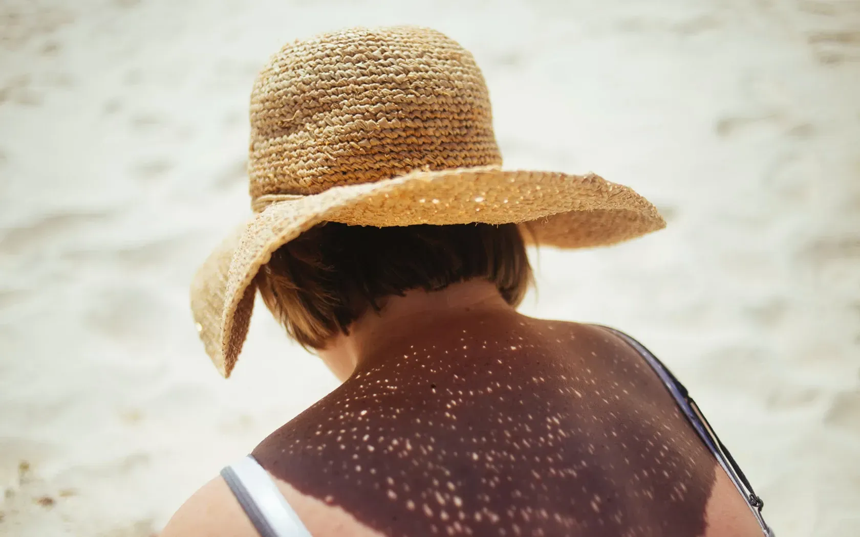 Person wearing a straw hat on a sunny beach, back to the camera, with light filtering through the hat.