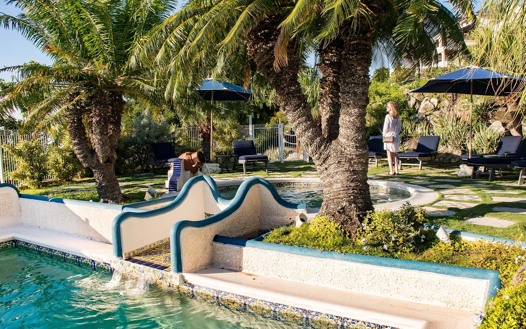 Poolside scene with palm trees, blue umbrellas, and a person standing nearby.