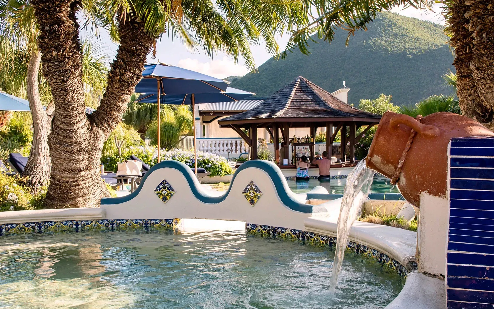 Pool with water feature, bar, palm trees, and mountain backdrop.