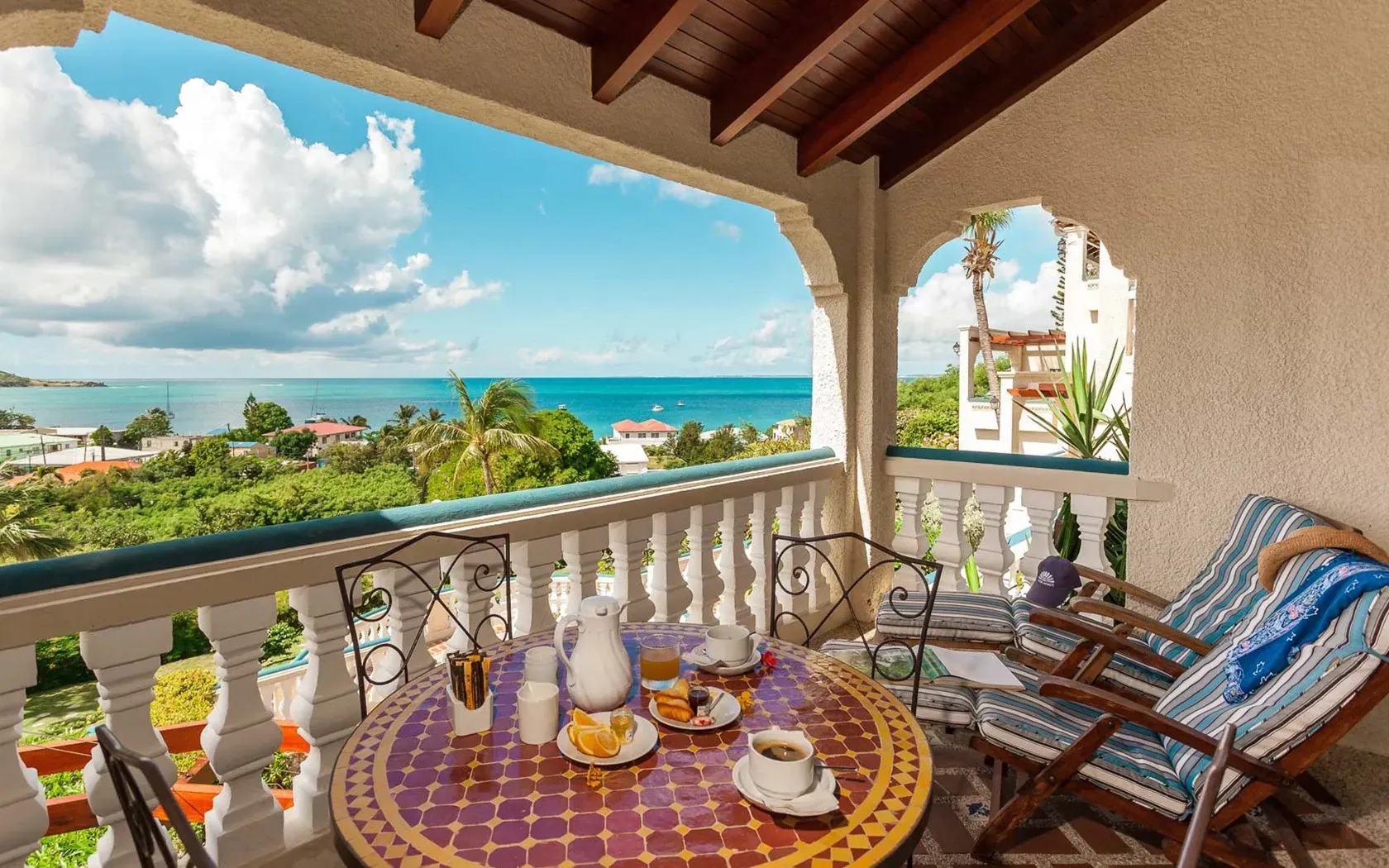 Balcony with breakfast table overlooking a tropical ocean view under a bright blue sky.