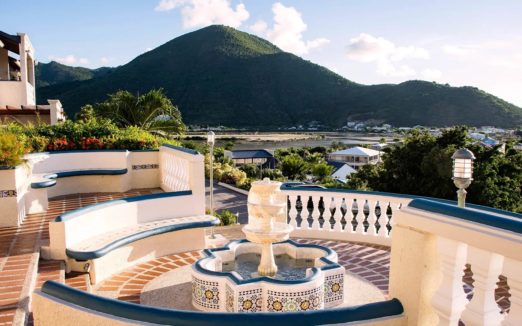Terrace with a fountain and benches, overlooking a green mountain and coastal town.