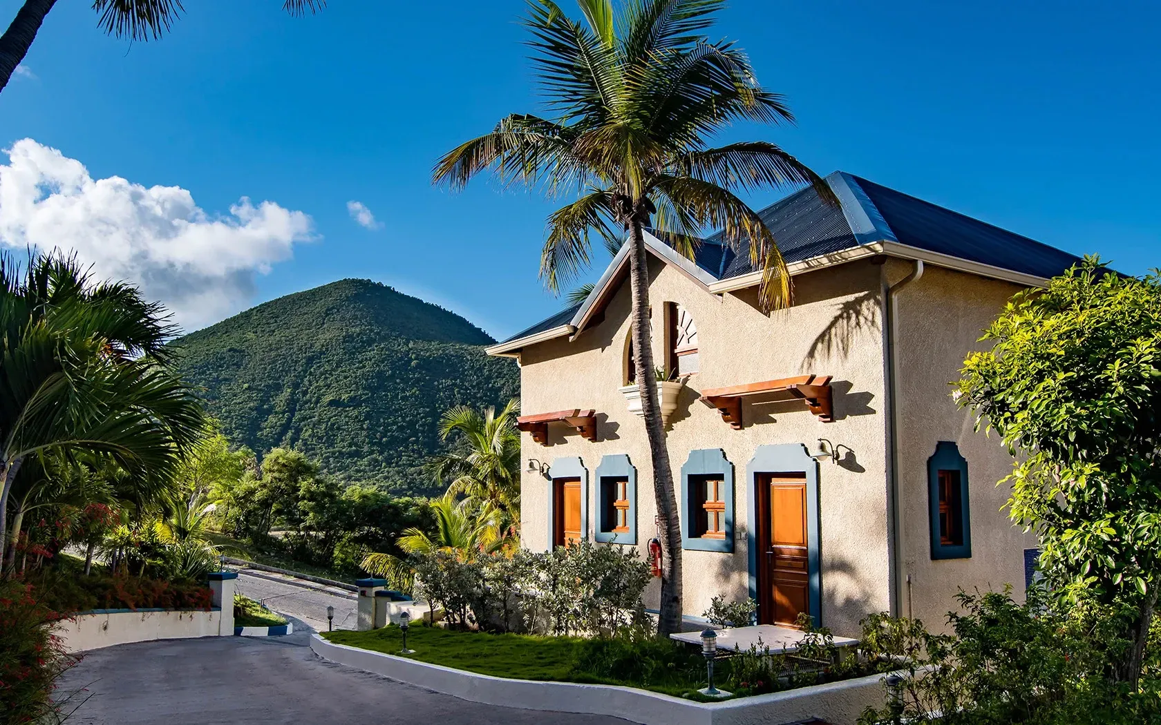 Tan house with blue roof, orange doors, surrounded by green trees and mountains under a blue sky.