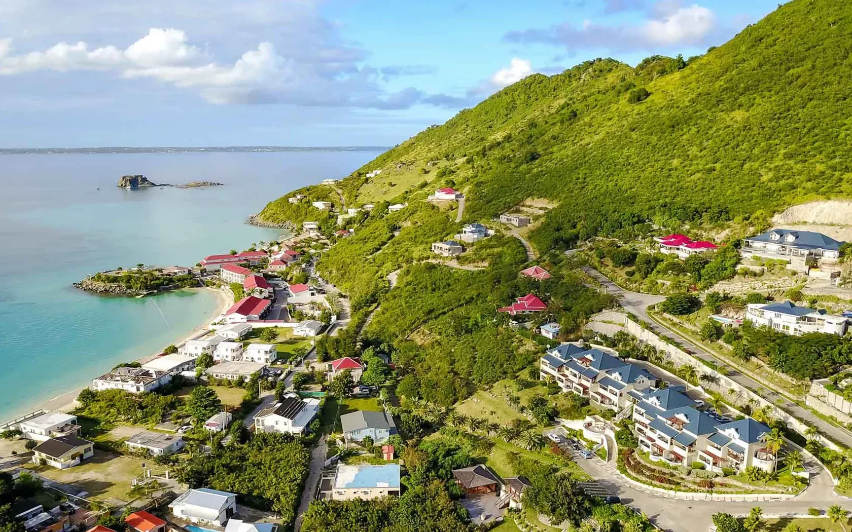 Coastal village with colorful buildings nestled at the foot of a lush green hillside, by turquoise waters.