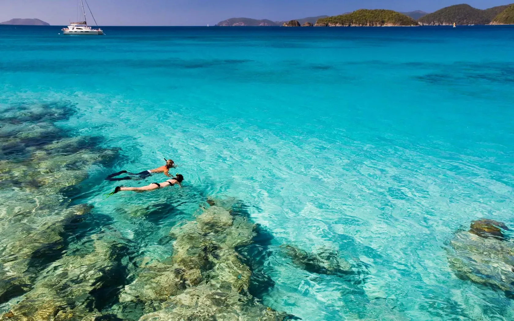 Two people snorkeling in clear, turquoise water near coral reefs, sailboat and islands in the background.