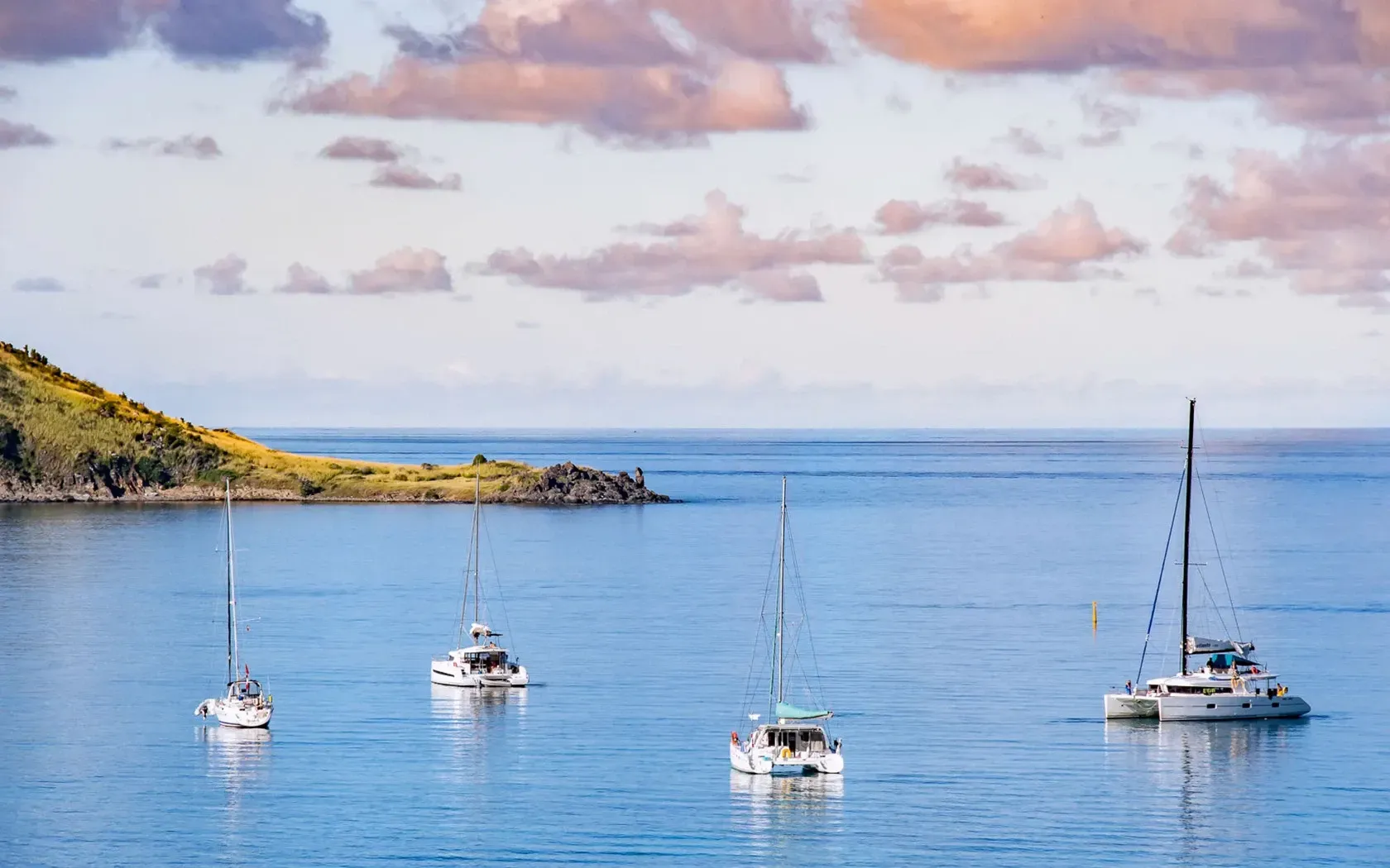 Sailboats anchored in calm blue water near a green island under a pastel sky.