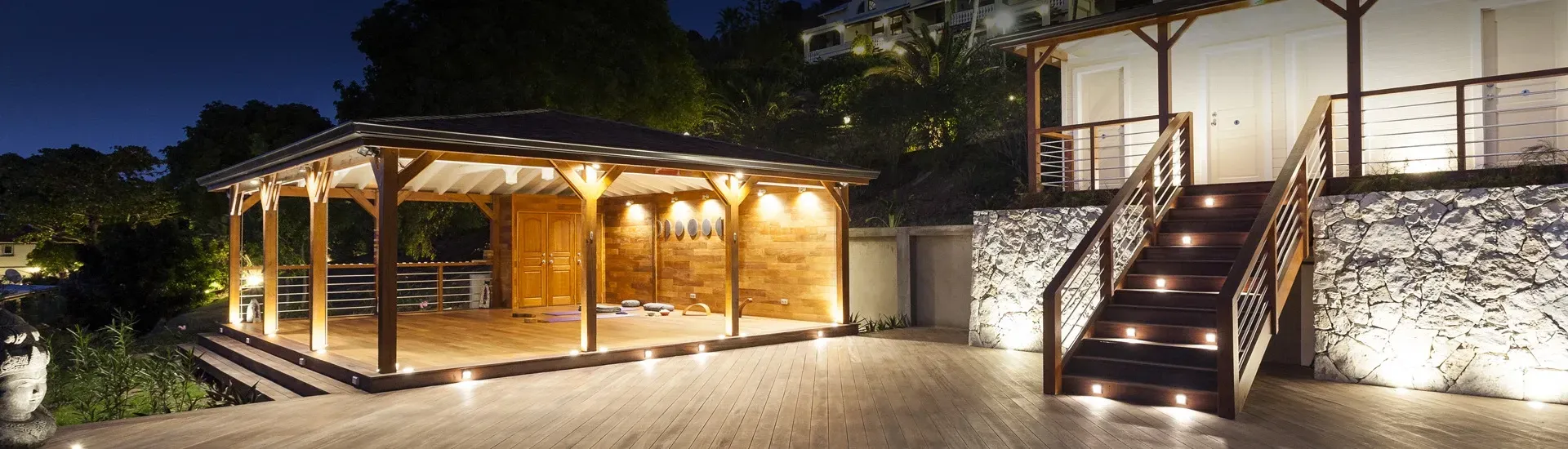 Lit outdoor wooden structures at night; stone walls, stairs, and landscaping.