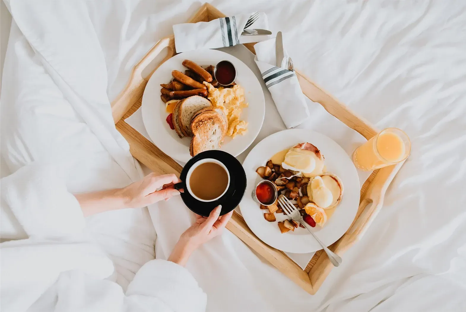 Breakfast in bed: Eggs, sausage, toast, and coffee on a tray, with person holding coffee in bed.
