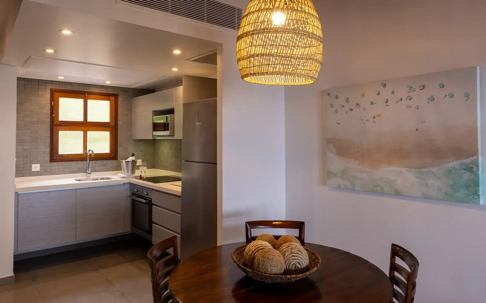 Kitchen and dining area with a table, chairs, and woven pendant light. Gray cabinets, and a seascape painting.