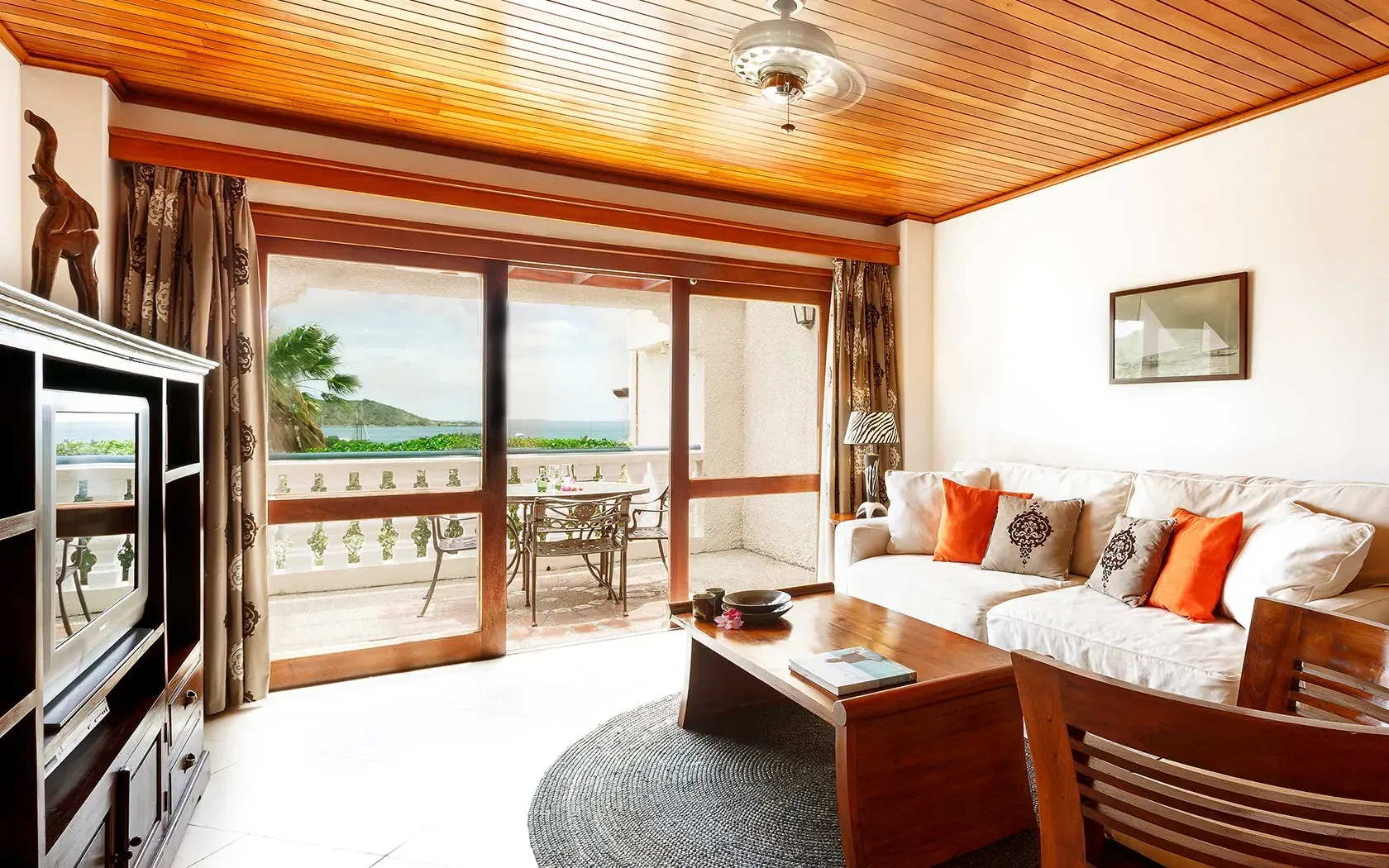 Living room with balcony overlooking a tropical view; wooden furniture, beige sofa, and wood-paneled ceiling.