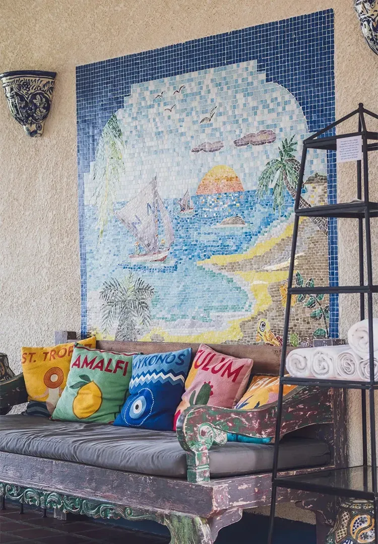 Sofa with colorful pillows in front of mosaic beach scene, with shelves and wall sconce.