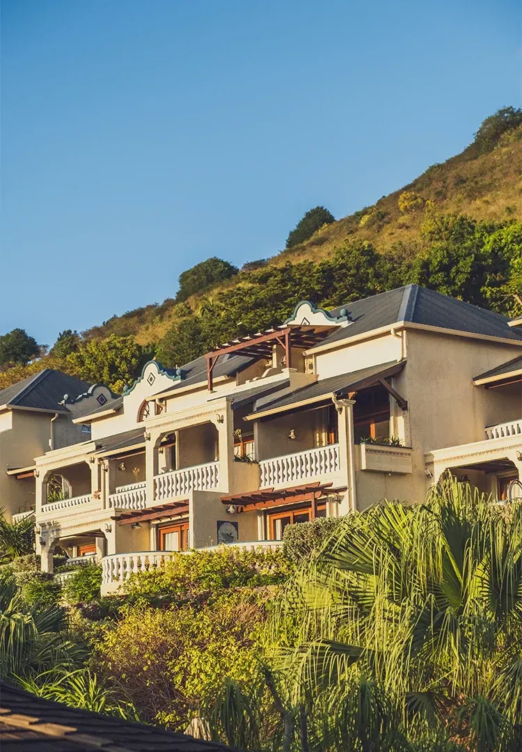 Beige multi-story buildings with balconies nestled against a green hillside under a blue sky.
