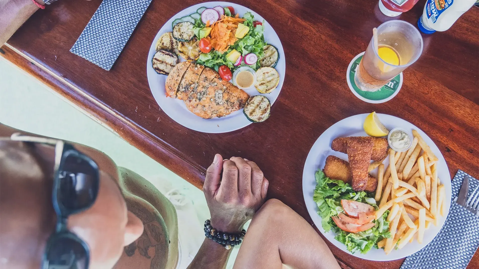 Person at a table eating grilled food and fish and chips, with a drink.
