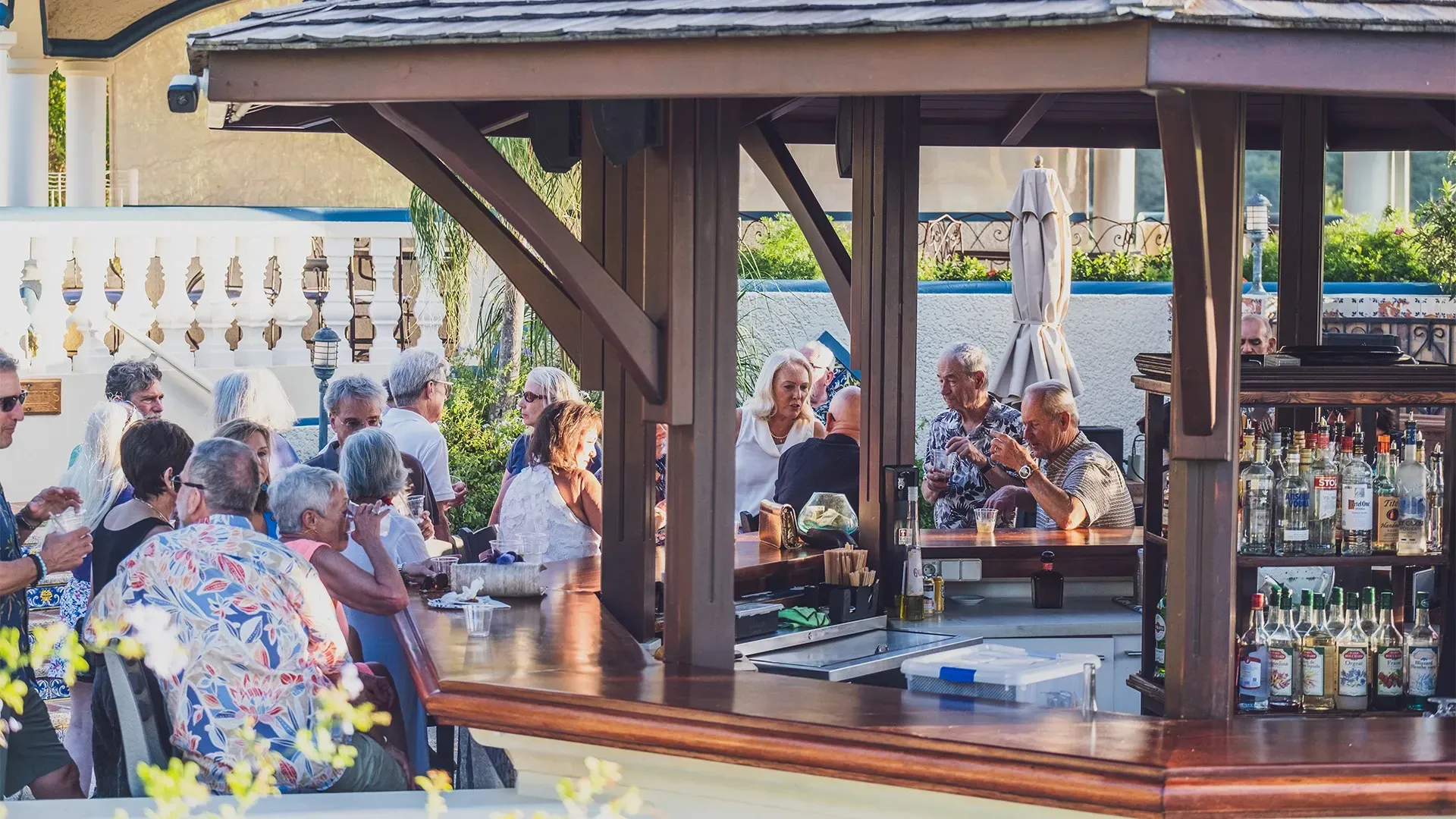 Outdoor bar with a gathering of people, some seated, others standing, enjoying drinks.