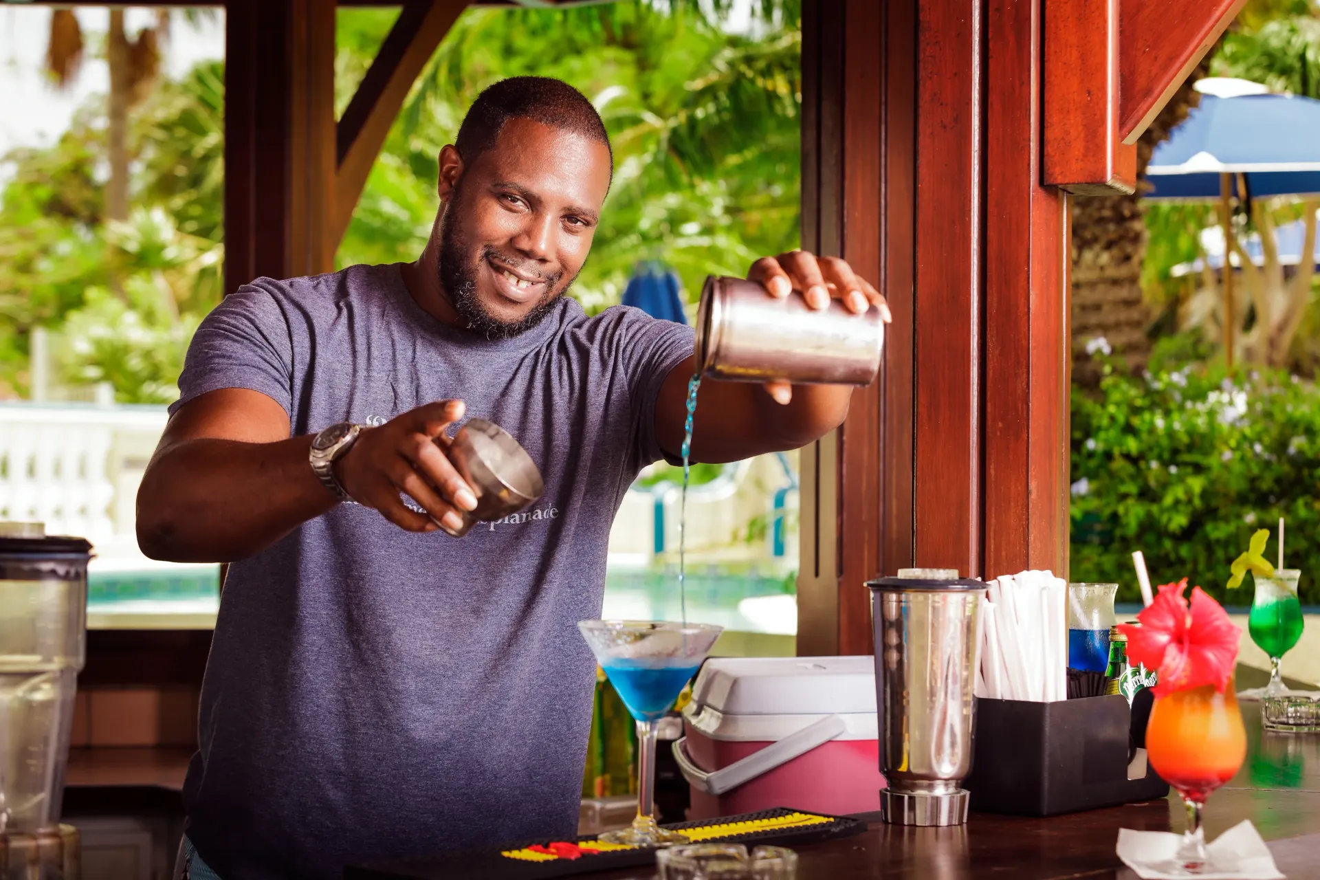 Bartender shaking a cocktail, smiles at the camera, outdoor bar with blue pool in background.