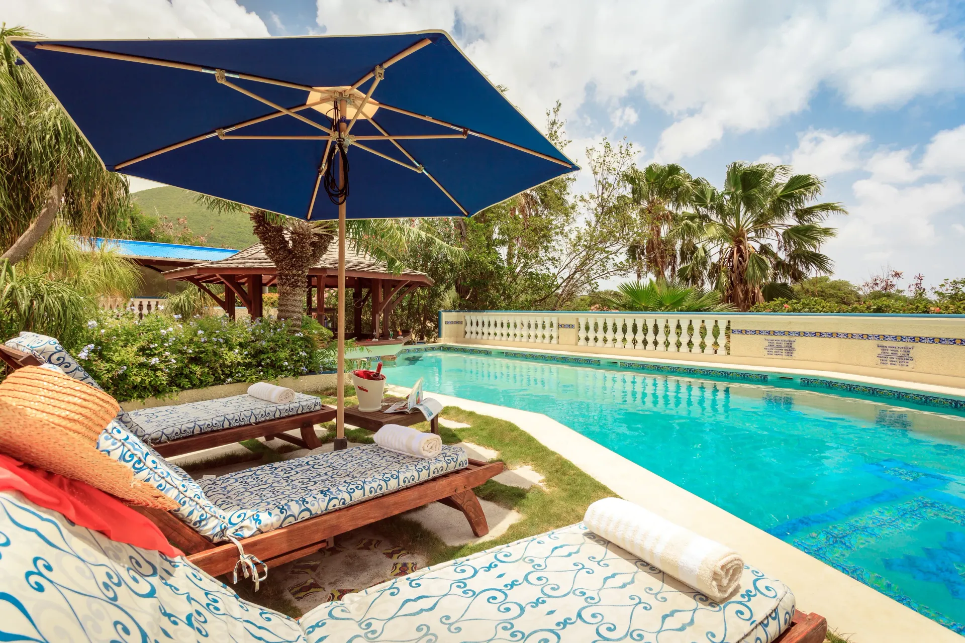 Lounge chairs by a pool with blue umbrella, sunny day. Lush greenery and a view.