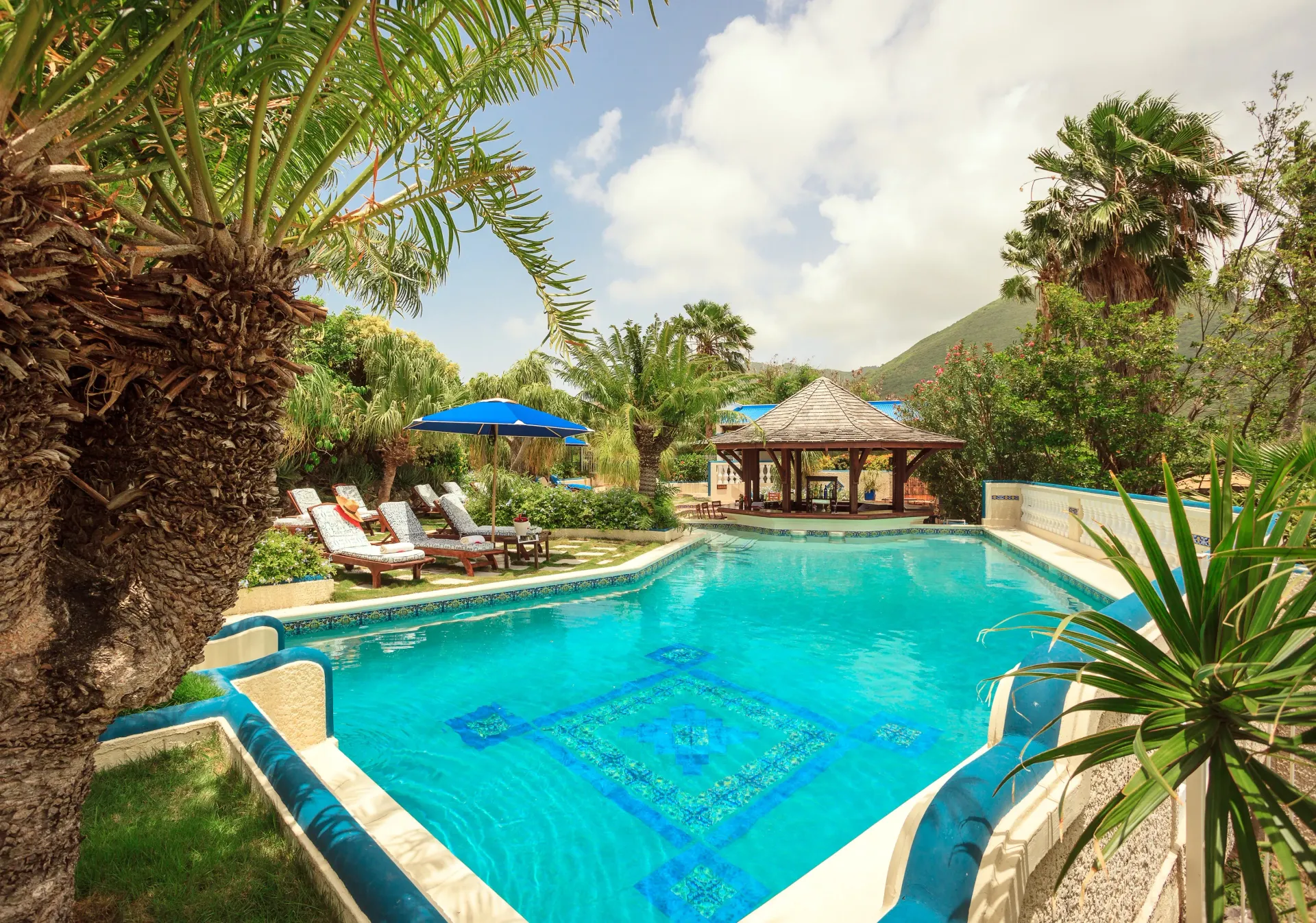 Pool with blue mosaic tile, lounge chairs, gazebo, and lush tropical greenery under a partly cloudy sky.
