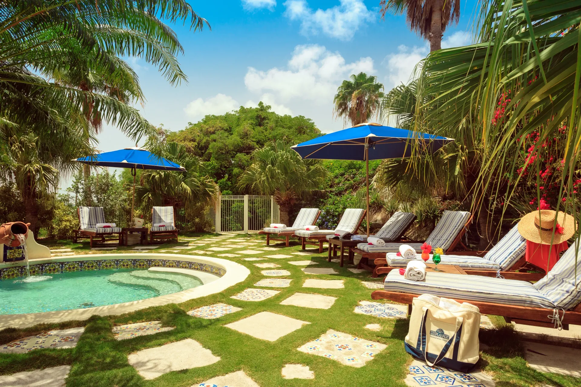 Poolside lounge area with chaise lounges, blue umbrellas, and a hot tub surrounded by lush greenery.