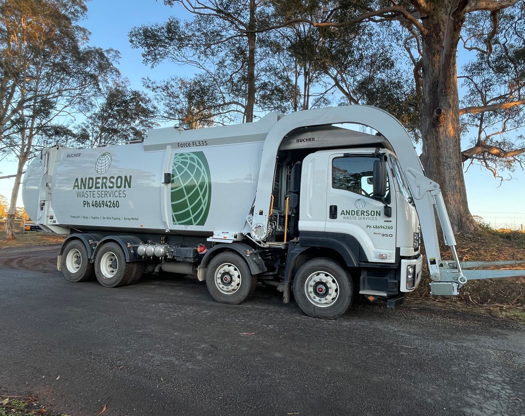 White garbage truck with a large lifting arm, parked outdoors near a tree.