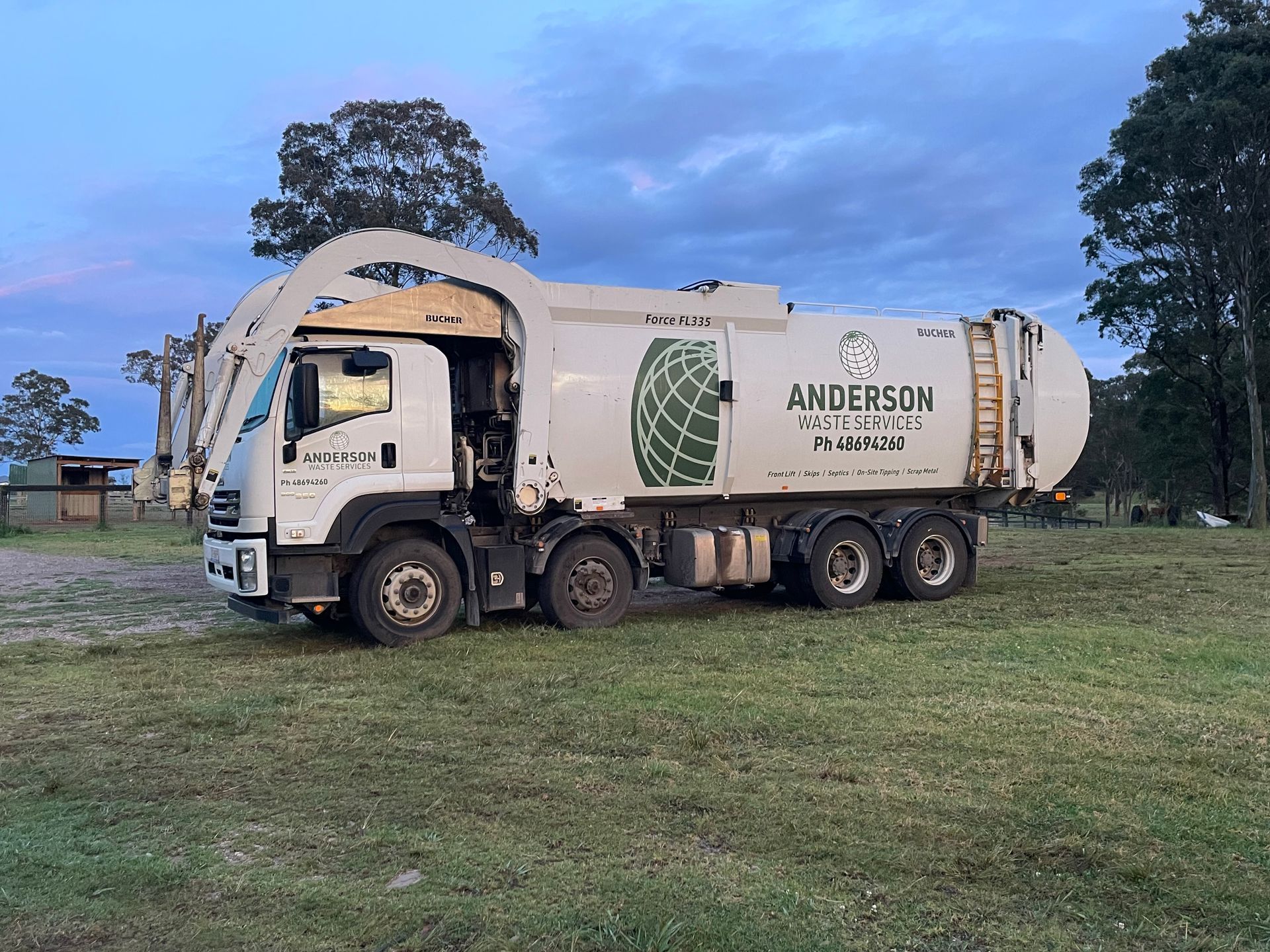 White Anderson waste truck on grass in front of trees, under a cloudy sky.
