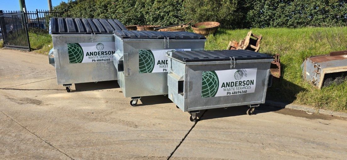 Three silver dumpsters with black lids on a paved area, green grass and foliage in the background.