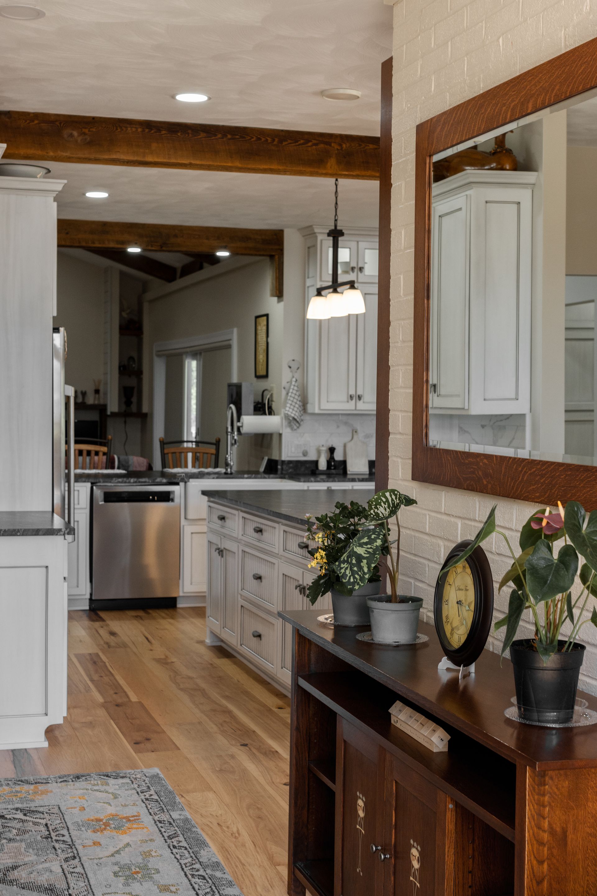 View of a kitchen with light wood floors, white cabinets, and dark countertops. Wooden beams and a mirror are also visible.