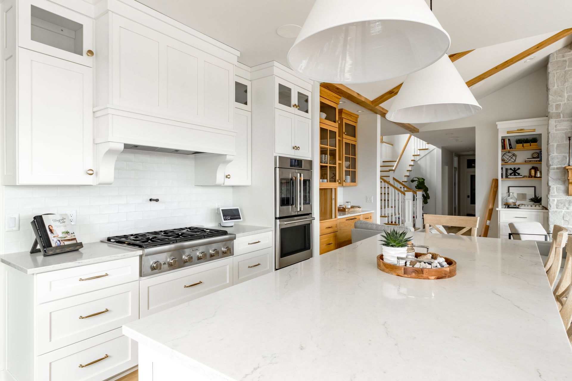 Modern white kitchen with island, range, and hanging lights. Wooden beams and staircase visible in the background.