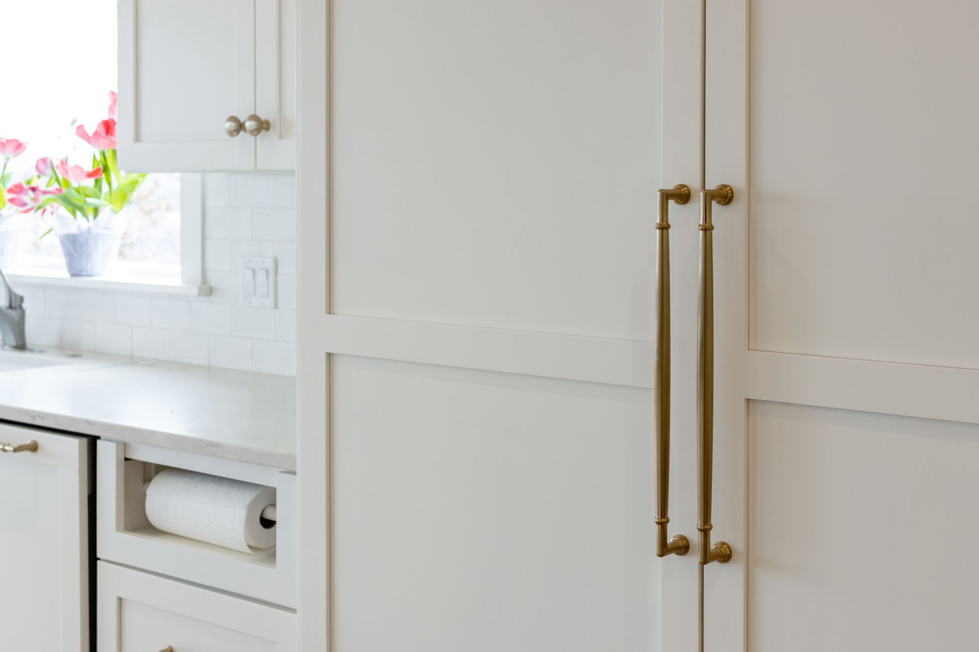White kitchen cabinets with brass handles, built-in paper towel holder, and a window with pink tulips.