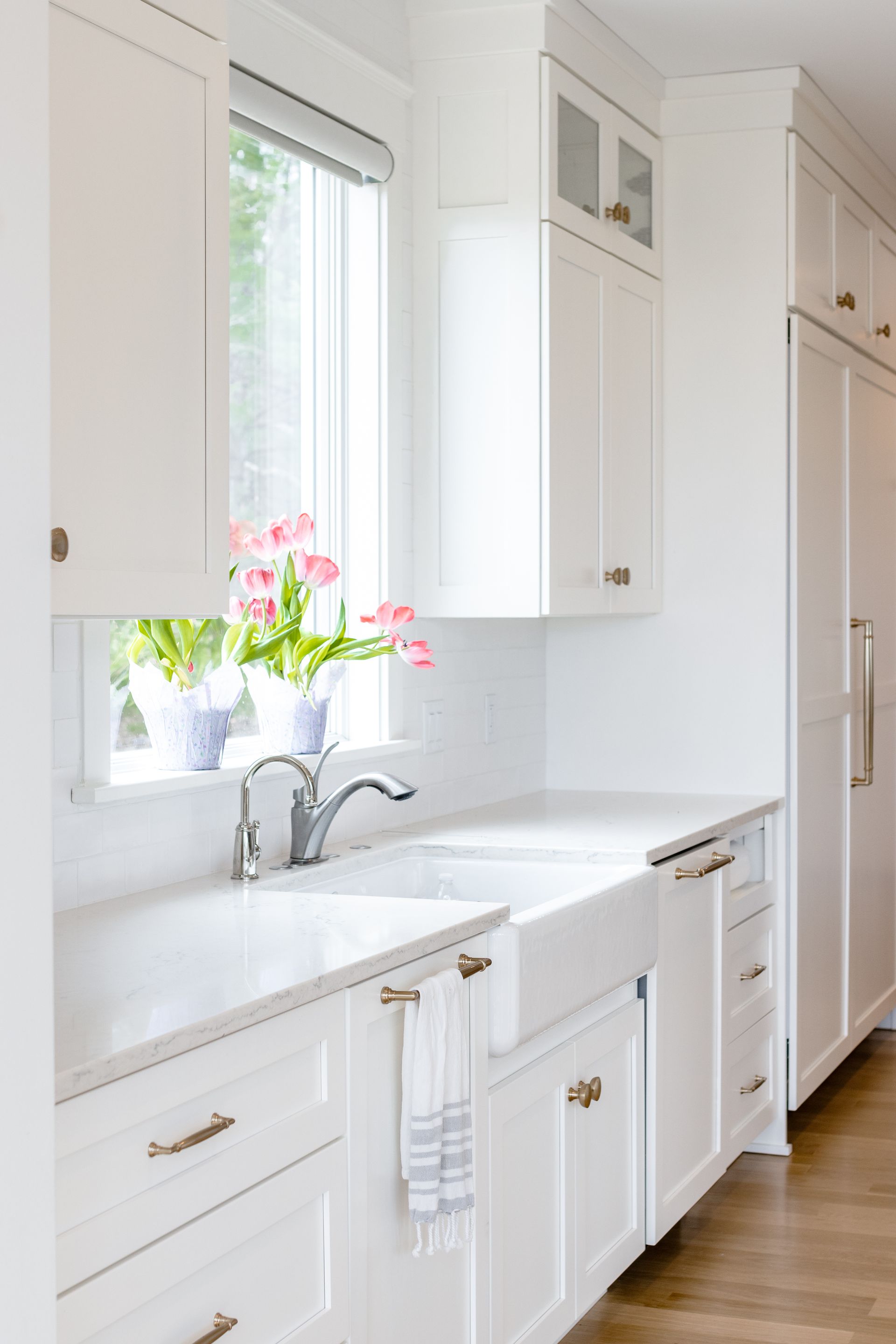 White kitchen with farmhouse sink, cabinets, window with pink tulips, gold hardware.