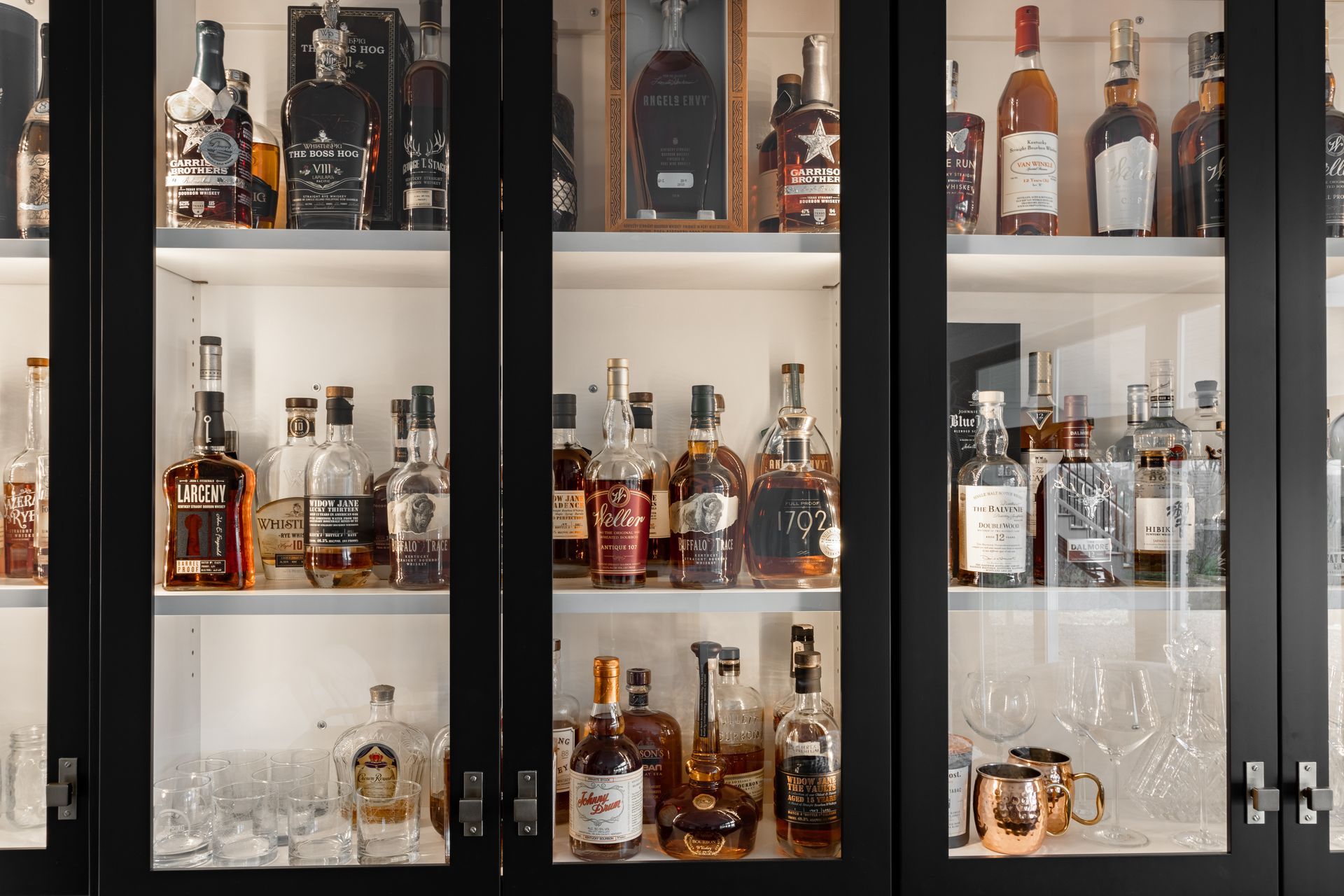 Liquor bottles on display in a black cabinet with glass doors. Shelves are illuminated; some empty glassware sits below.