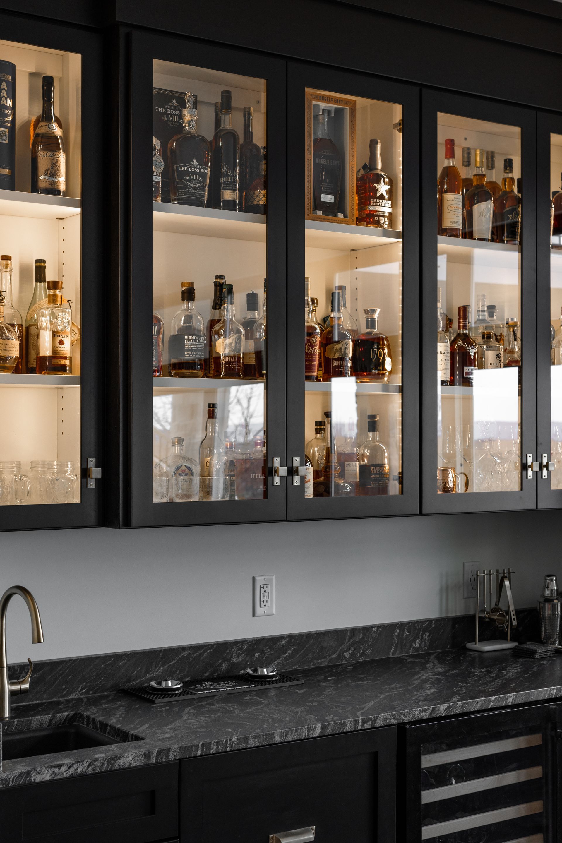 Black cabinets with glass doors, backlit to illuminate liquor bottles, above a dark counter and sink.