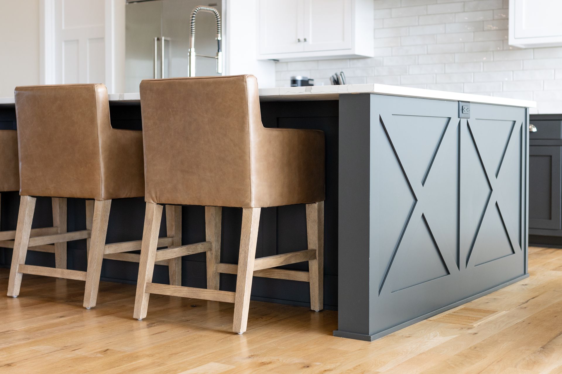 Kitchen island with gray base, wooden X design, light-colored wooden floors, and tan bar stools.