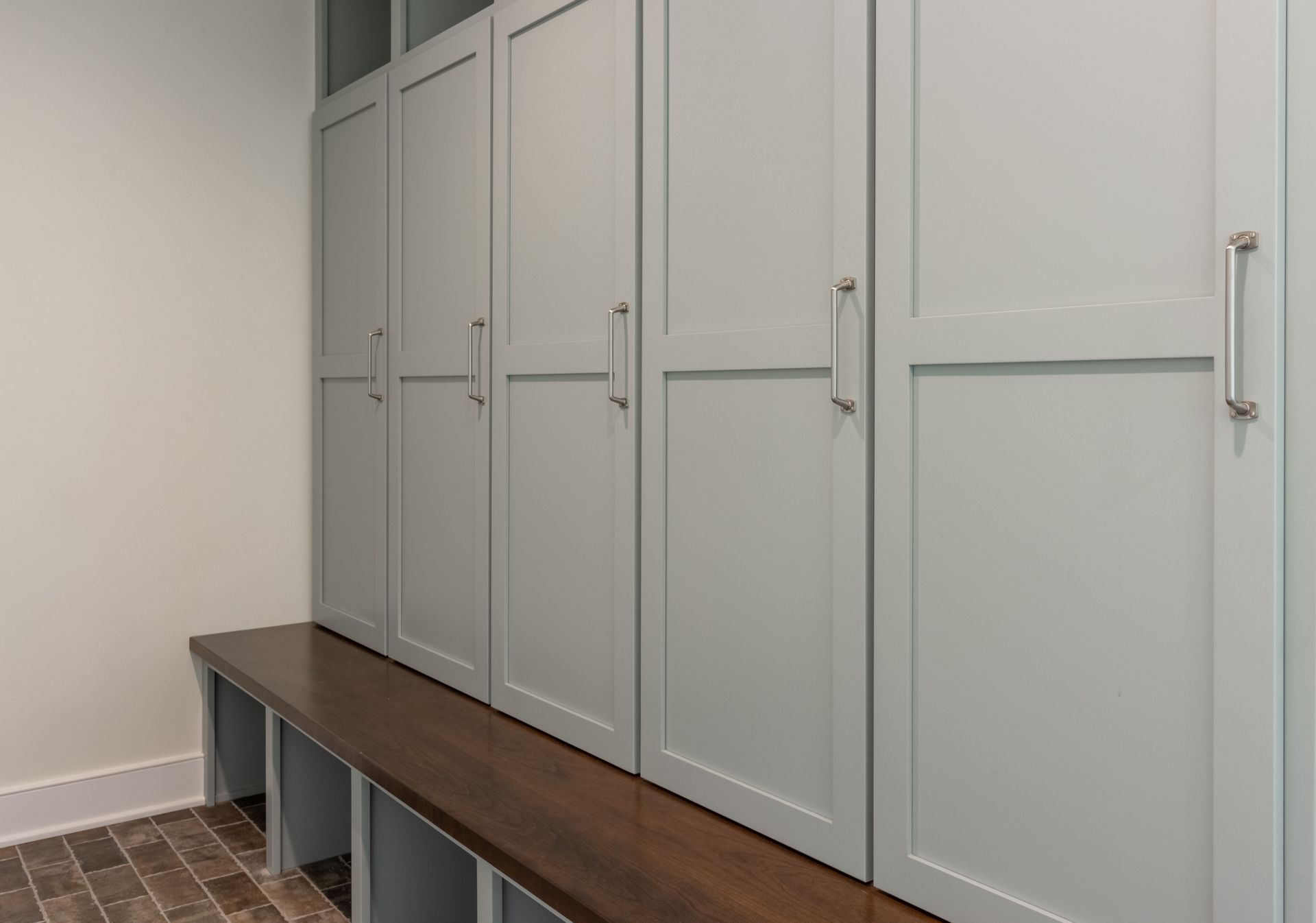 Gray storage lockers with silver handles above a brown bench in a room.