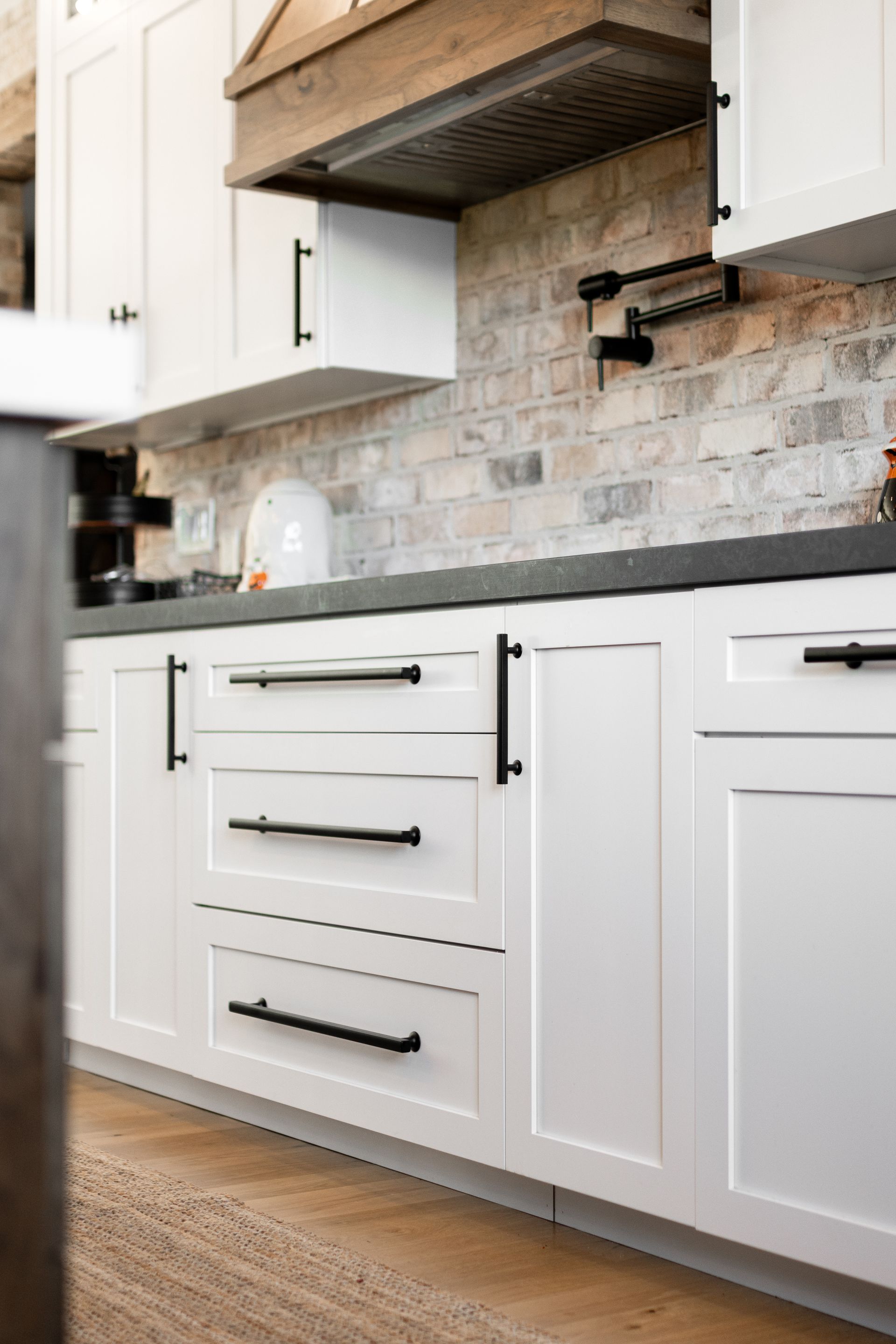 White kitchen cabinets with black hardware against a brick backsplash, beneath a wooden hood.