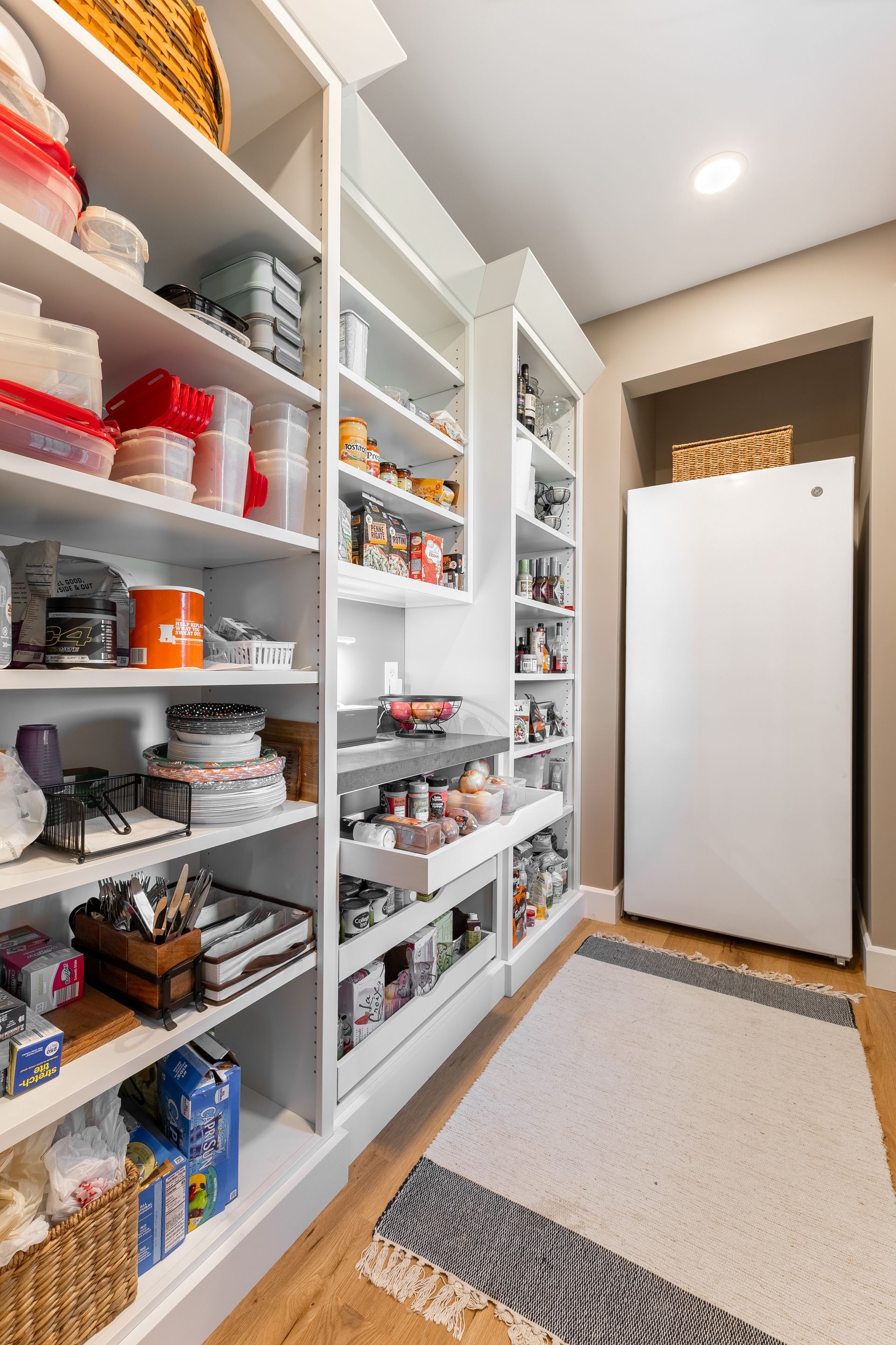 Walk-in pantry with white shelves filled with food items, pull-out drawers, and a refrigerator on a rug.