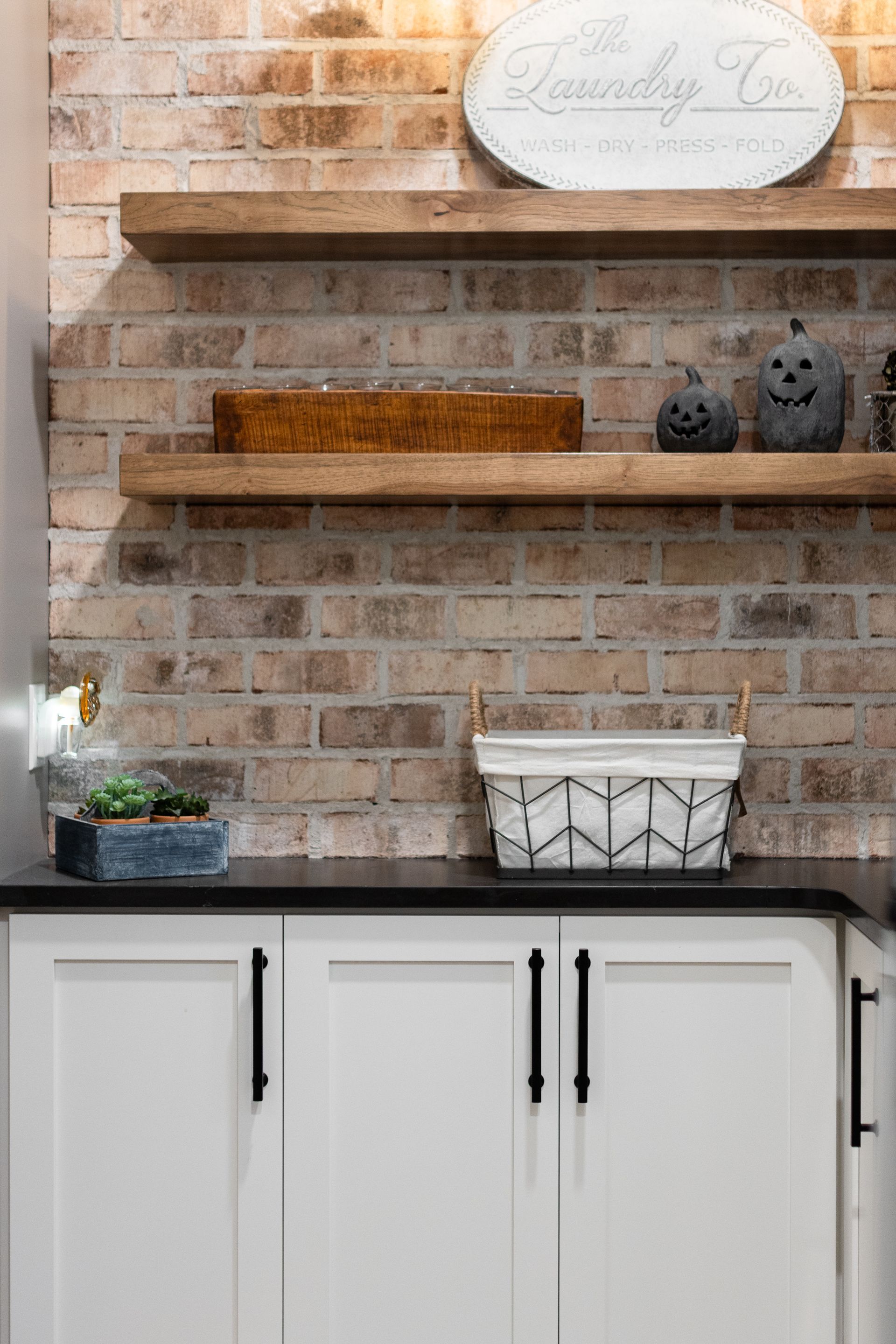 White cabinets with black handles, brick wall shelves with decorations, and a white basket.