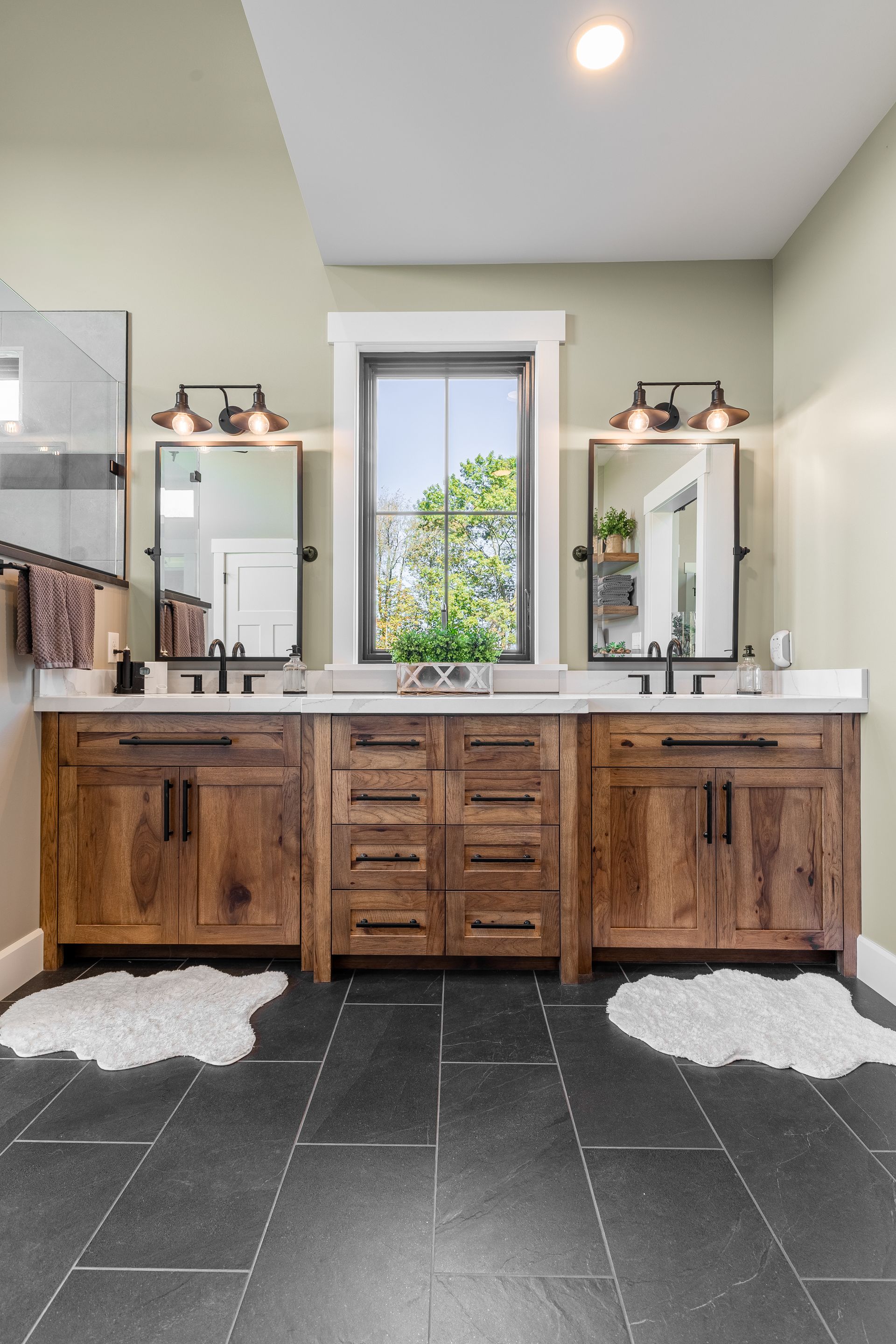 Bathroom with dark tile floor, wooden vanity, mirrors, and window with greenery.